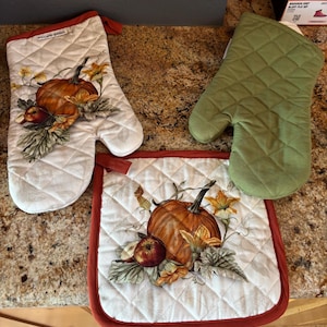 Puede incluir: Un juego de manopla y salvamanteles acolchados blancos con temática de cosecha de otoño. La manopla y el salvamanteles presentan un diseño de calabaza, manzana y flores. También se incluye un guante de cocina verde liso.