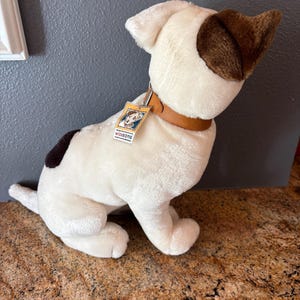 Puede incluir: Un perro de peluche blanco con orejas marrones y una mancha marr&oacute;n en la espalda. Tiene un collar marr&oacute;n y una etiqueta que dice "Wishbone". El perro est&aacute; sentado sobre una superficie marr&oacute;n.
