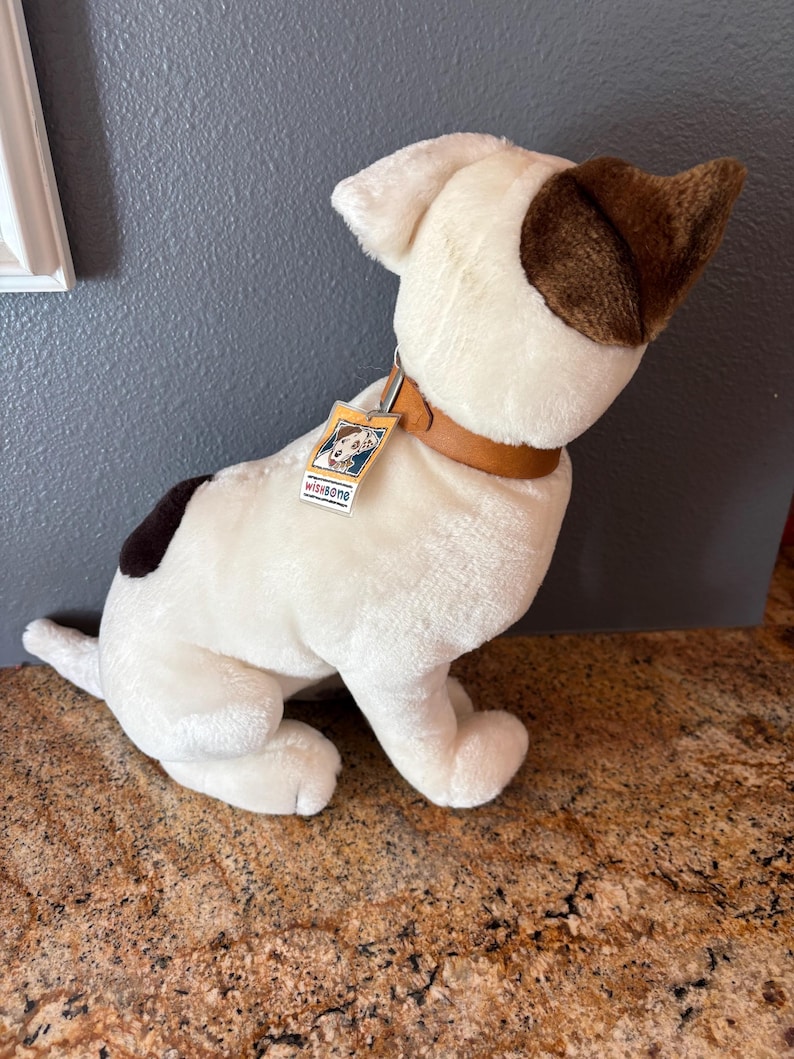 Puede incluir: Un perro de peluche blanco con orejas marrones y una mancha marr&oacute;n en la espalda. Tiene un collar marr&oacute;n y una etiqueta que dice "Wishbone". El perro est&aacute; sentado sobre una superficie marr&oacute;n.