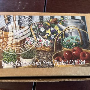May include: A cardboard box featuring a garden-themed illustration. The image depicts baskets, flowers, a watering can, and apples. Text on the box reads "Herb Marker and Seed Packet Gift Set."