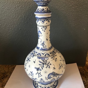 May include: A tall, white ceramic vase with intricate blue floral and bird designs. The vase has a bulbous body, a long neck, and a decorative top. The base and top are adorned with detailed patterns.