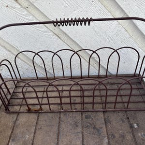 May include: A rectangular wire basket with a dark brown finish, featuring a grid-patterned base and arched wire sides. The basket includes a single tall, arched handle with a coiled wire grip at the top center for carrying and storage.