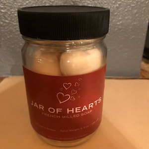 May include: A glass jar filled with white heart-shaped soap bars. The jar has a red label with the text "Jar of Hearts French Milled Soap" and "Total Weight 6.5 oz / 184 g".