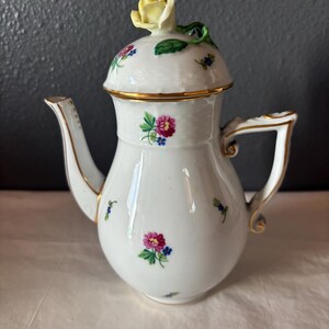 May include: White porcelain teapot with a yellow rose on the lid. The teapot has a gold rim and handle, and is decorated with pink, blue, and green floral designs. The spout is curved and the handle is a squared shape.
