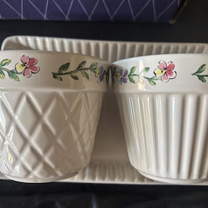 May include: Two off-white ceramic flower pots with a matching tray. One pot has a diamond pattern, the other has vertical lines. Both pots have a floral design around the rim with pink, yellow, and blue flowers.