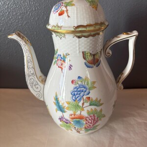 May include: White porcelain coffee pot with a lid, adorned with colorful floral and butterfly designs. The pot features a gold trim and handle, and a decorative spout. The lid is topped with a yellow floral accent.