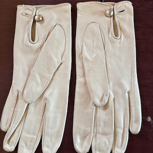 May include: A pair of off-white leather gloves featuring a smooth finish, a wrist-length cut, and a decorative silver-toned metal button closure at the top of each wrist. The gloves have a classic design with stitched seams along the fingers.