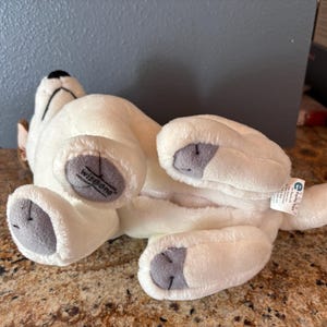 Puede incluir: Un juguete de peluche blanco con forma de perro tumbado boca arriba, con almohadillas grises y una nariz negra. El juguete tiene la palabra "Wisdom" impresa en una pata. Una peque&ntilde;a etiqueta est&aacute; sujeta a la cola del perro.