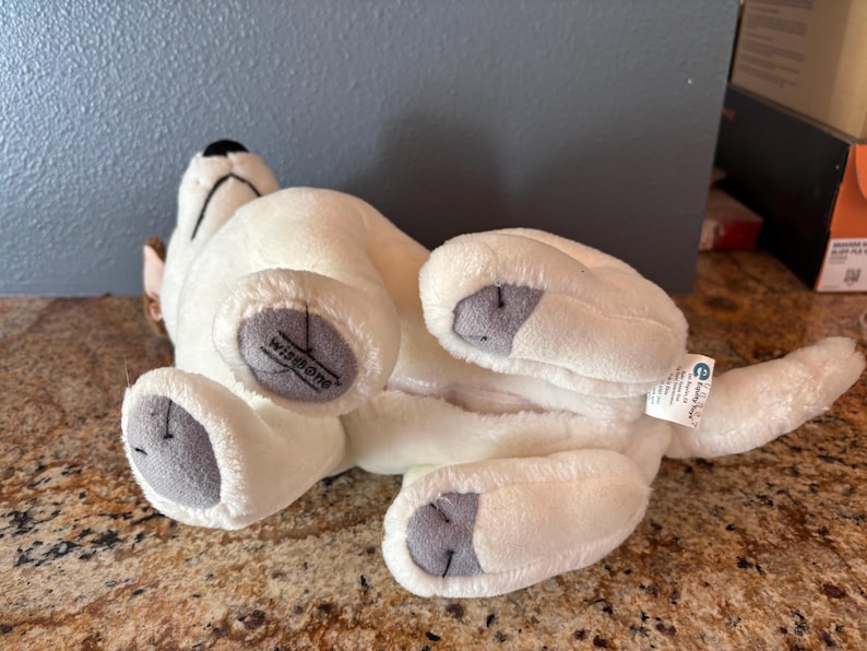 Puede incluir: Un juguete de peluche blanco con forma de perro tumbado boca arriba, con almohadillas grises y una nariz negra. El juguete tiene la palabra "Wisdom" impresa en una pata. Una peque&ntilde;a etiqueta est&aacute; sujeta a la cola del perro.