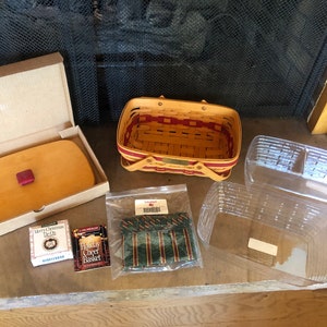 May include: A collection of Longaberger items, including a rectangular basket with red accents, a wooden lid with a red knob, and clear plastic inserts. Also included are two small boxes and a package of green and red ribbon.