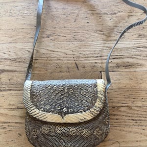 May include: A vintage crossbody bag with a snakeskin pattern in shades of gray and tan. The bag features a flap closure with a decorative trim and a gray shoulder strap. The bag is made of a textured material.