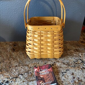 May include: A light brown woven basket with a handle, and a small book titled "Classic Baskets" by Longaberger. The basket is rectangular with a woven design and a wooden handle. The book has a red cover with a picture of a basket.