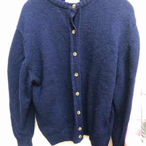 May include: A navy blue cardigan sweater with a round neckline and button-up closure. The sweater is made of a thick knit material and has long sleeves. The buttons are a light brown color. The sweater is hanging on a wooden hanger.
