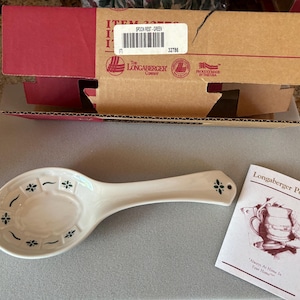 May include: A white ceramic spoon rest with green floral accents, a Longaberger Pottery product. The spoon rest is oval-shaped with a handle and a small hole. A box and a product information card are also in the image.