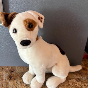 Puede incluir: Un peluche de perro con cuerpo blanco, orejas marrones y manchas marrones alrededor de los ojos y en la espalda. El perro tiene una nariz negra y est&aacute; sentado.