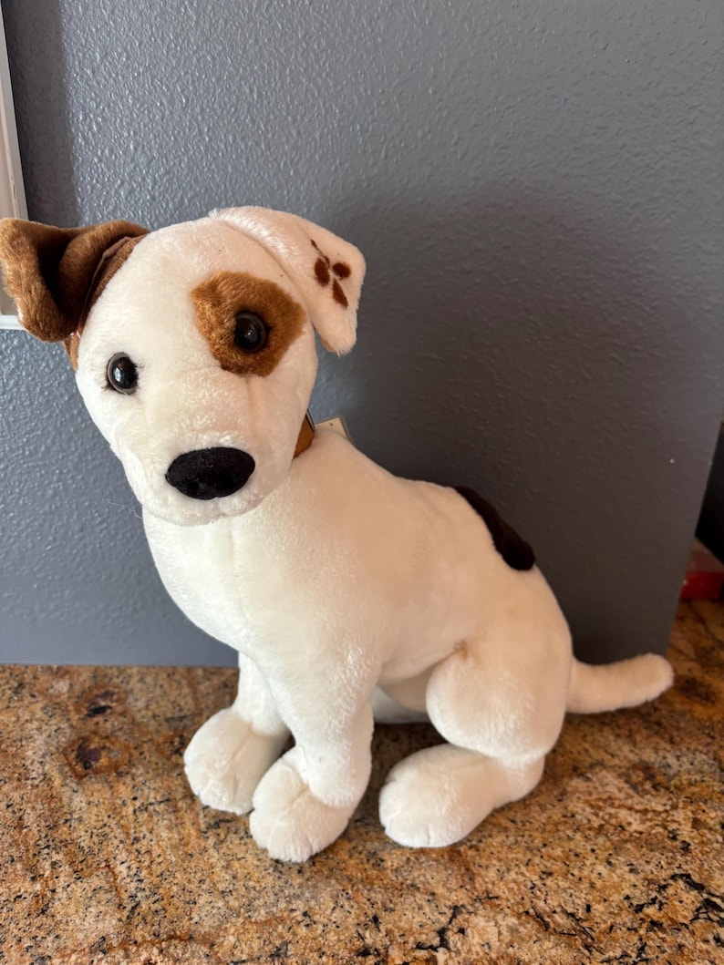 Puede incluir: Un peluche de perro con cuerpo blanco, orejas marrones y manchas marrones alrededor de los ojos y en la espalda. El perro tiene una nariz negra y est&aacute; sentado.