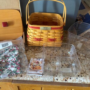 May include: A woven basket with a handle, red accents, and a Longaberger label. Includes a wooden lid with a red knob, a plastic insert, and a Christmas-themed fabric liner.