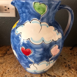 May include: A blue ceramic pitcher with a white interior and a handle. The pitcher is decorated with white cloud shapes and colorful heart shapes in red, green, and pink. The pitcher is sitting on a brown surface.
