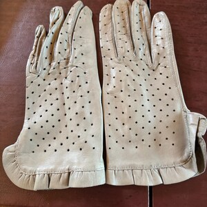 May include: A pair of beige leather gloves featuring a perforated design across the back of the hands and fingers, finished with a ruffled edge at the wrist. The gloves are constructed with visible stitching and a smooth, light-colored leather finish.