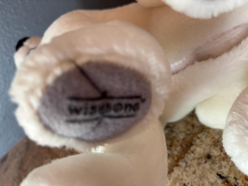 Puede incluir: Primer plano del pie de un juguete de peluche blanco con una almohadilla gris. La almohadilla tiene la palabra "wisdone" impresa en negro. El juguete es suave y esponjoso, con un cuerpo color crema.