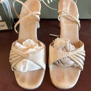 May include: A pair of vintage beige high-heeled sandals. The sandals feature crisscross straps across the foot and ankle straps. The heels are a light beige color. The sandals are labeled "Rangeri Made in Florence Italy".
