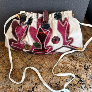 May include: White shoulder bag with a decorative design in shades of burgundy, red, and green. The bag features a long, thin white strap and a gold-colored clasp. The design incorporates various textures and patterns.