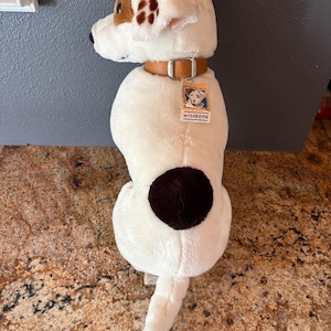 Puede incluir: Un juguete de peluche con forma de perro blanco con manchas marrones y un collar de cuero marr&oacute;n. El perro tiene una oreja marr&oacute;n y una cola. Una etiqueta con el texto "wishbone" est&aacute; adjunta al collar.