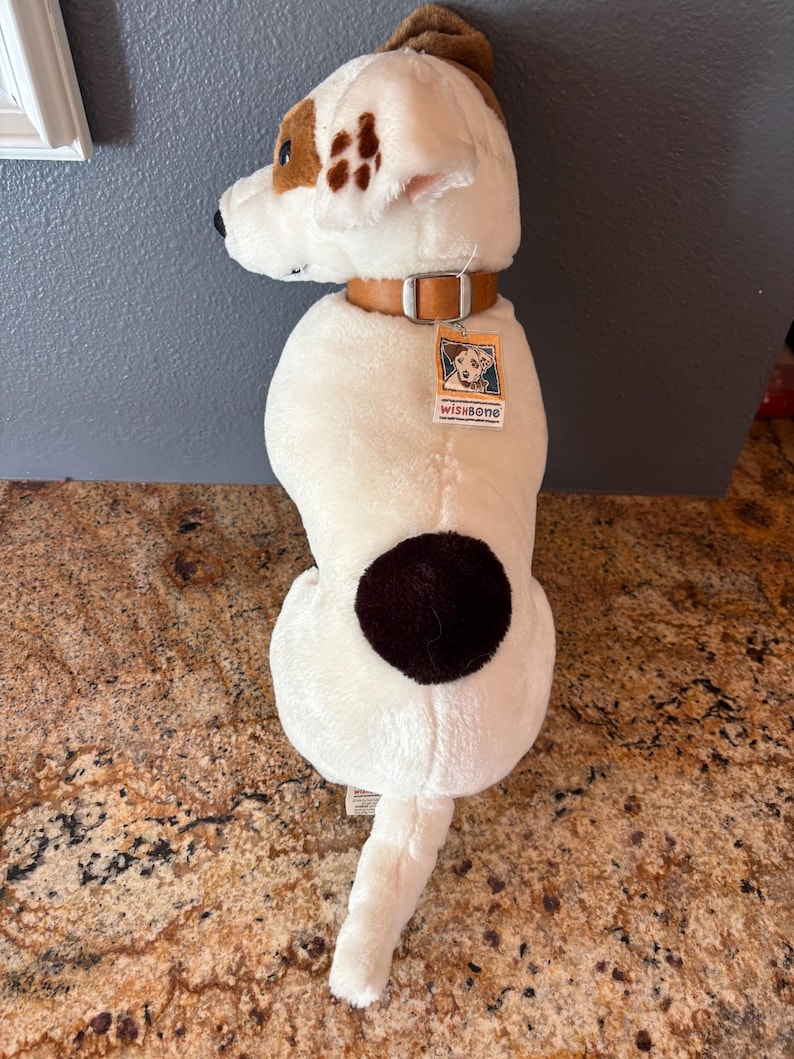 Puede incluir: Un juguete de peluche con forma de perro blanco con manchas marrones y un collar de cuero marr&oacute;n. El perro tiene una oreja marr&oacute;n y una cola. Una etiqueta con el texto "wishbone" est&aacute; adjunta al collar.