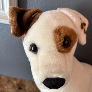 Puede incluir: Un perro de peluche, predominantemente blanco con manchas marrones alrededor de los ojos y las orejas. El perro tiene una nariz negra y ojos oscuros, lo que le da un aspecto amigable. El juguete est&aacute; dise&ntilde;ado para parecerse a un Jack Russell Terrier.