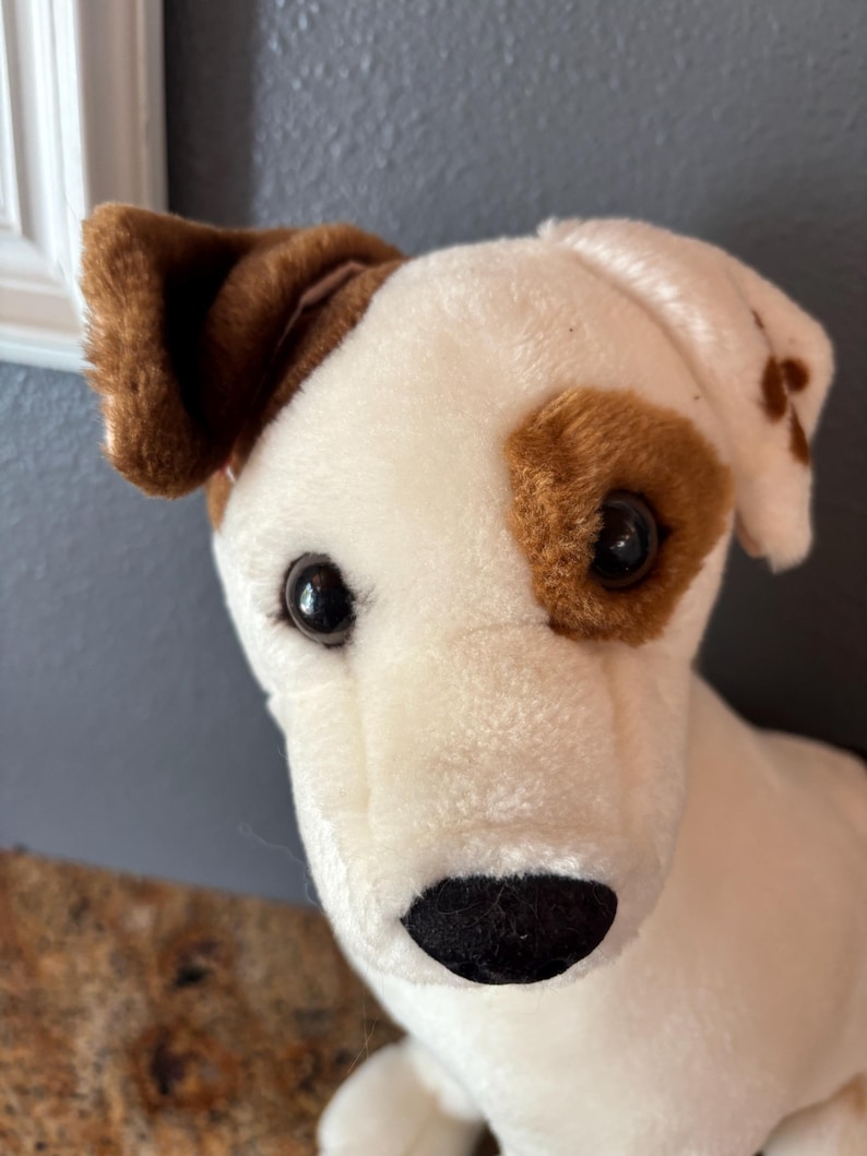Puede incluir: Un perro de peluche, predominantemente blanco con manchas marrones alrededor de los ojos y las orejas. El perro tiene una nariz negra y ojos oscuros, lo que le da un aspecto amigable. El juguete est&aacute; dise&ntilde;ado para parecerse a un Jack Russell Terrier.