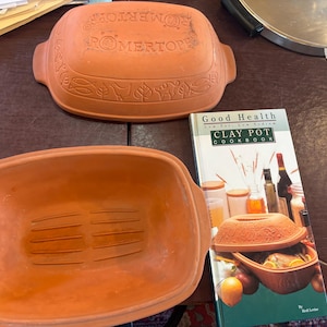 May include: A terracotta Romertopf clay baker with a lid featuring embossed animal and leaf motifs, accompanied by a Good Health Low Fat, Low Sodium Clay Pot Cookbook. The rectangular baker has integrated handles and a ribbed interior base.