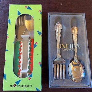 May include: Two sets of children's silverware. One set is in a green and red box with a Christmas tree design and the text "Mary Engelbreit". The other set is in a silver box with the text "ONEIDA SILVERPLATE".