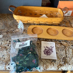 May include: A Longaberger Baskets brand wooden tray with three round indentations. The tray is made of light brown wood and is sitting on a light brown countertop. There is a small bag of green and black fabric scraps, a clear plastic container, and a light brown wicker basket in the background.