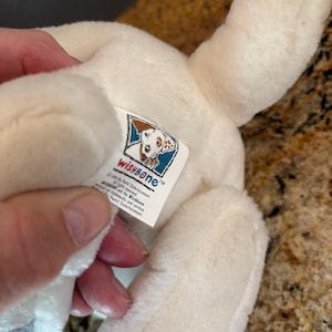 Puede incluir: Un juguete de peluche blanco con forma de perro con manchas negras en las patas y una etiqueta que dice "WISHBONE". El juguete es suave y esponjoso, con un dise&ntilde;o detallado. La etiqueta presenta una ilustraci&oacute;n de perro de dibujos animados.