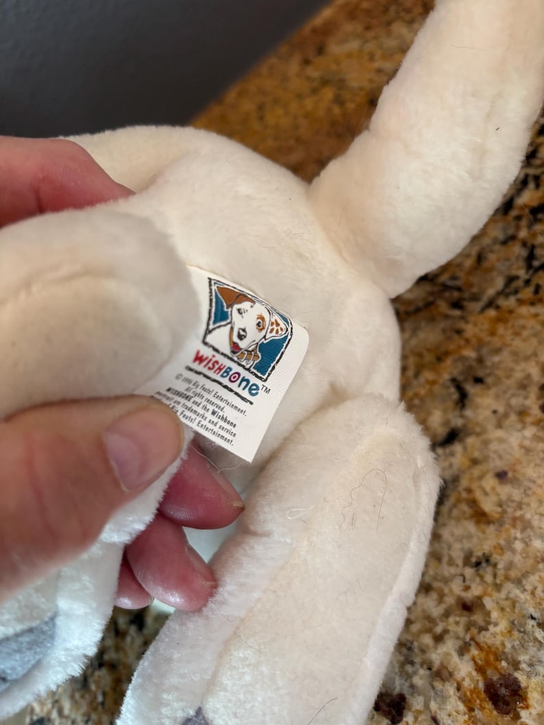 Puede incluir: Un juguete de peluche blanco con forma de perro con manchas negras en las patas y una etiqueta que dice "WISHBONE". El juguete es suave y esponjoso, con un dise&ntilde;o detallado. La etiqueta presenta una ilustraci&oacute;n de perro de dibujos animados.
