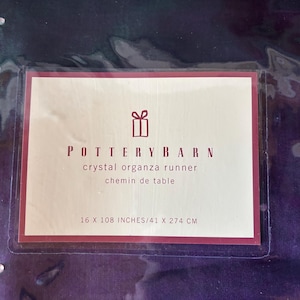 May include: A crystal organza runner from Pottery Barn. The label reads "crystal organza runner chemin de table" and measures 16 x 108 inches (41 x 274 cm). The runner is a deep purple color.