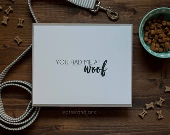 May include: A rectangular white card with a gray border featuring the black text "YOU HAD ME AT woof" centered on the front. Below the text, the small gray handle "@otterandlane" is printed. The card is surrounded by dog treats and a leash.