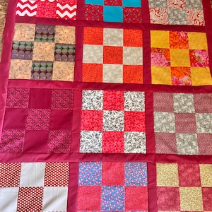May include: A patchwork quilt with a variety of patterned squares in shades of red, pink, yellow, and blue. The squares are arranged in a grid pattern and separated by a solid red border. The quilt features floral, geometric, and abstract designs.