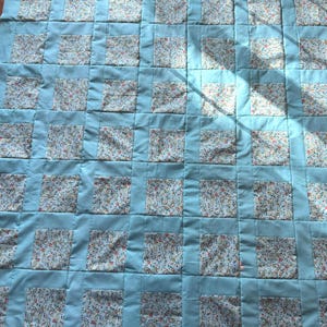 May include: A handmade quilt with a light blue background and a patchwork design. The quilt features squares of floral fabric framed by light blue borders. The overall pattern is a grid of alternating floral and solid squares.
