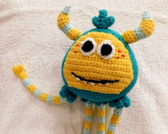 Op de afbeelding: Een gehaakt geel en blauw monster speelgoed met lange gestreepte benen en een vriendelijk gezicht.