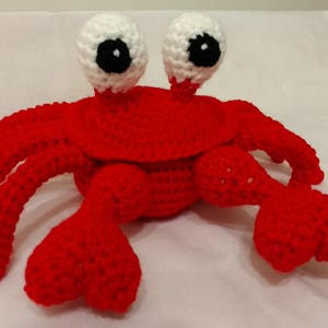 May include: A red crocheted crab with white eyes and black pupils. The crab has eight legs and is sitting on a white surface.