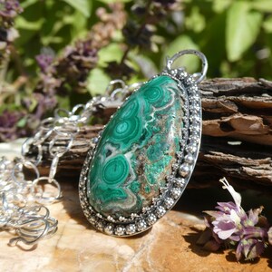 May include: A silver pendant necklace featuring a large, oval malachite stone with swirling green patterns. The stone is set in a detailed silver bezel and hangs from a silver chain. The background includes wood and foliage.