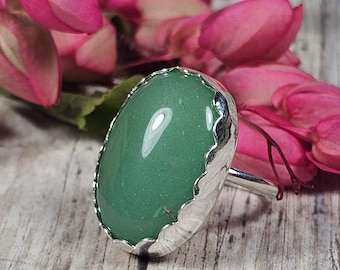 May include: A silver ring featuring a large, oval-shaped, green gemstone. The gemstone is set in a decorative, scalloped bezel. Pink flowers are in the background, and the ring is displayed on a light-colored wooden surface.