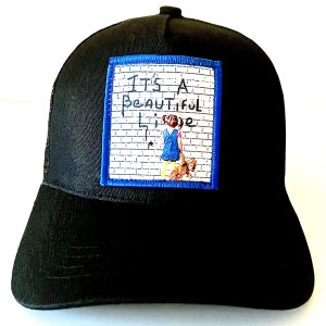 以下が含まれることがあります： 青い縁取りの白地に「It's a Beautiful Life」と書いてあるパッチがついた黒いトラッカーハット。レンガの壁に女の子と犬の絵が描かれている。