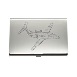 May include: A silver business card holder featuring an etched outline of a private jet. The jet's wings, tail, and fuselage are clearly defined. The card holder has a shiny, reflective surface. The design is a detailed illustration.