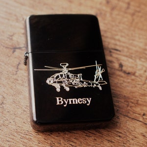 May include: A black lighter featuring a silver helicopter design and the name "Byrnesy". The rectangular lighter has a glossy finish. The background is a wooden surface. The lighter is a classic design.