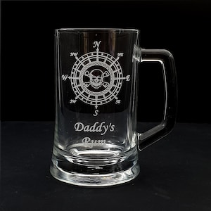 Könnte beinhalten: Ein klares Glasbierkrug mit einem Kompass- und Totenkopfdesign, das in die Vorderseite geätzt ist. Der Krug hat einen Henkel und der Text "Daddy's Rum" ist unterhalb des Designs eingeätzt.