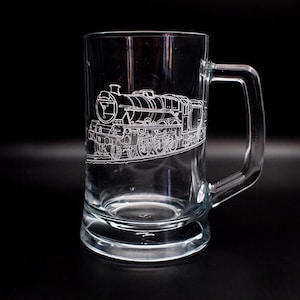 May include: A clear glass beer mug featuring an etched design of a vintage steam locomotive. The mug has a handle and a rounded base.