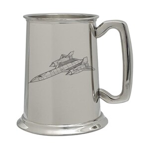 May include: A polished silver-toned metal beer mug featuring a detailed black line art engraving of an SR-71 Blackbird aircraft on the front. The mug has a cylindrical body, a wide base, and a sturdy, curved handle on the right side.