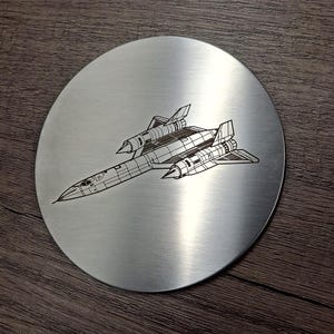 May include: A circular brushed metal coaster featuring a black line art engraving of a supersonic reconnaissance aircraft. The design depicts the distinct silhouette of the jet with its elongated fuselage, dual engine nacelles, and swept-back wings.